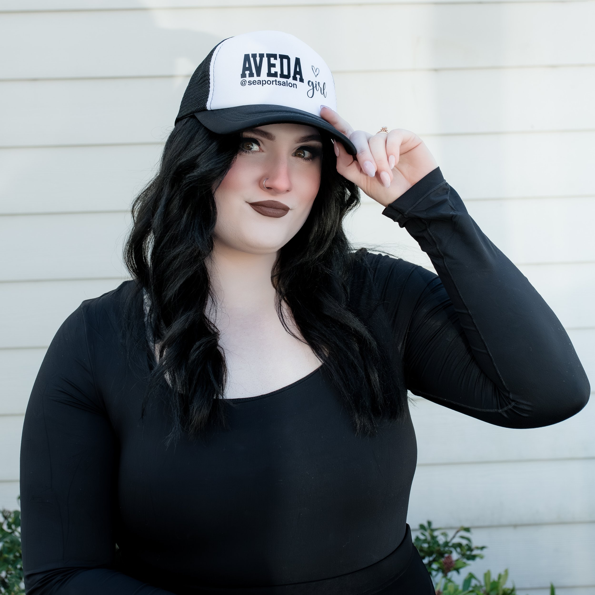 Woman with dark hair wearing a black long-sleeve top and a trucker hat with the words "AVEDA girl" on it, standing in front of a white wooden wall.