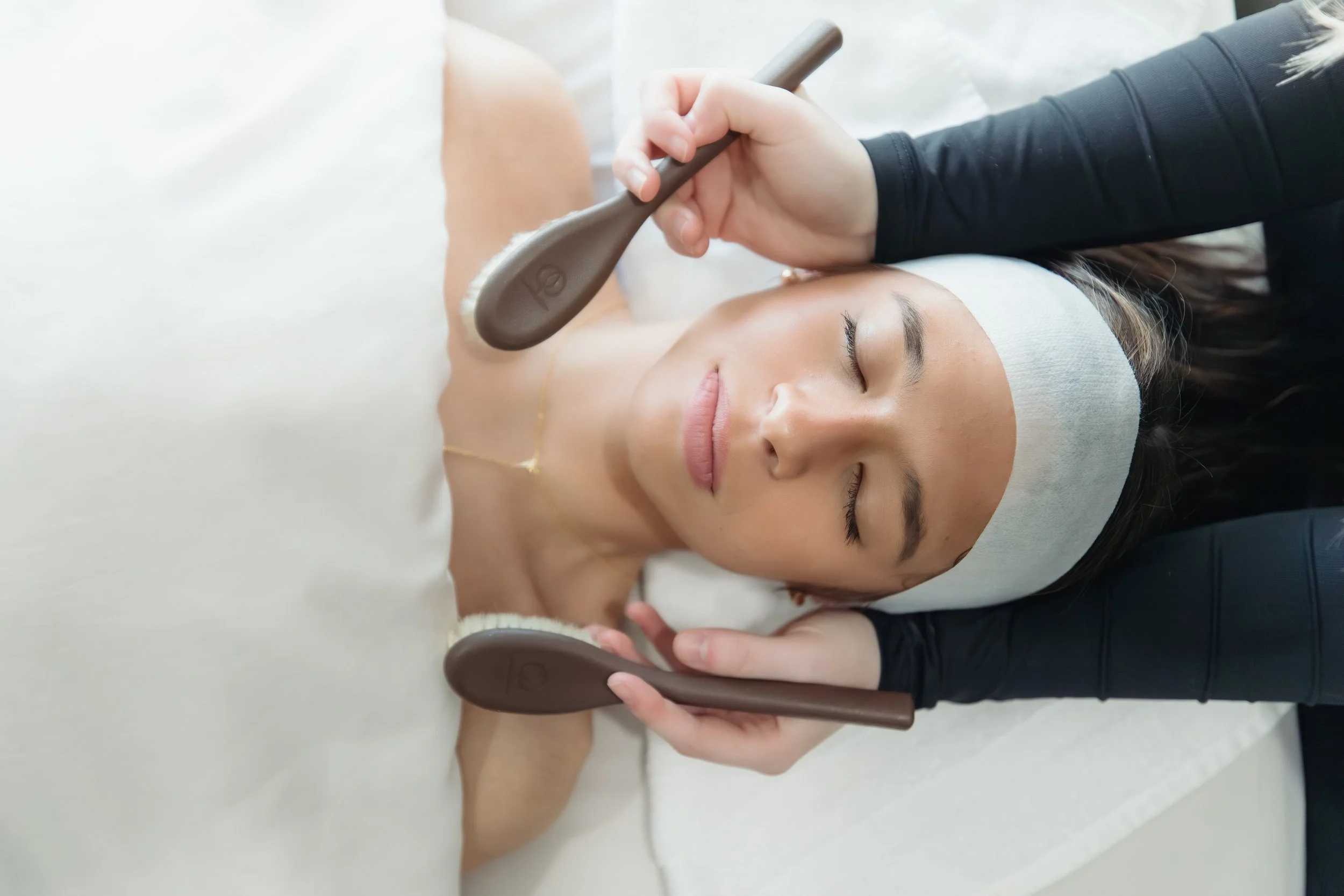 A woman receiving a facial treatment while lying down with her eyes closed, a headband around her forehead, and a massage therapist applying a facial massage with two brown facial massage tools.