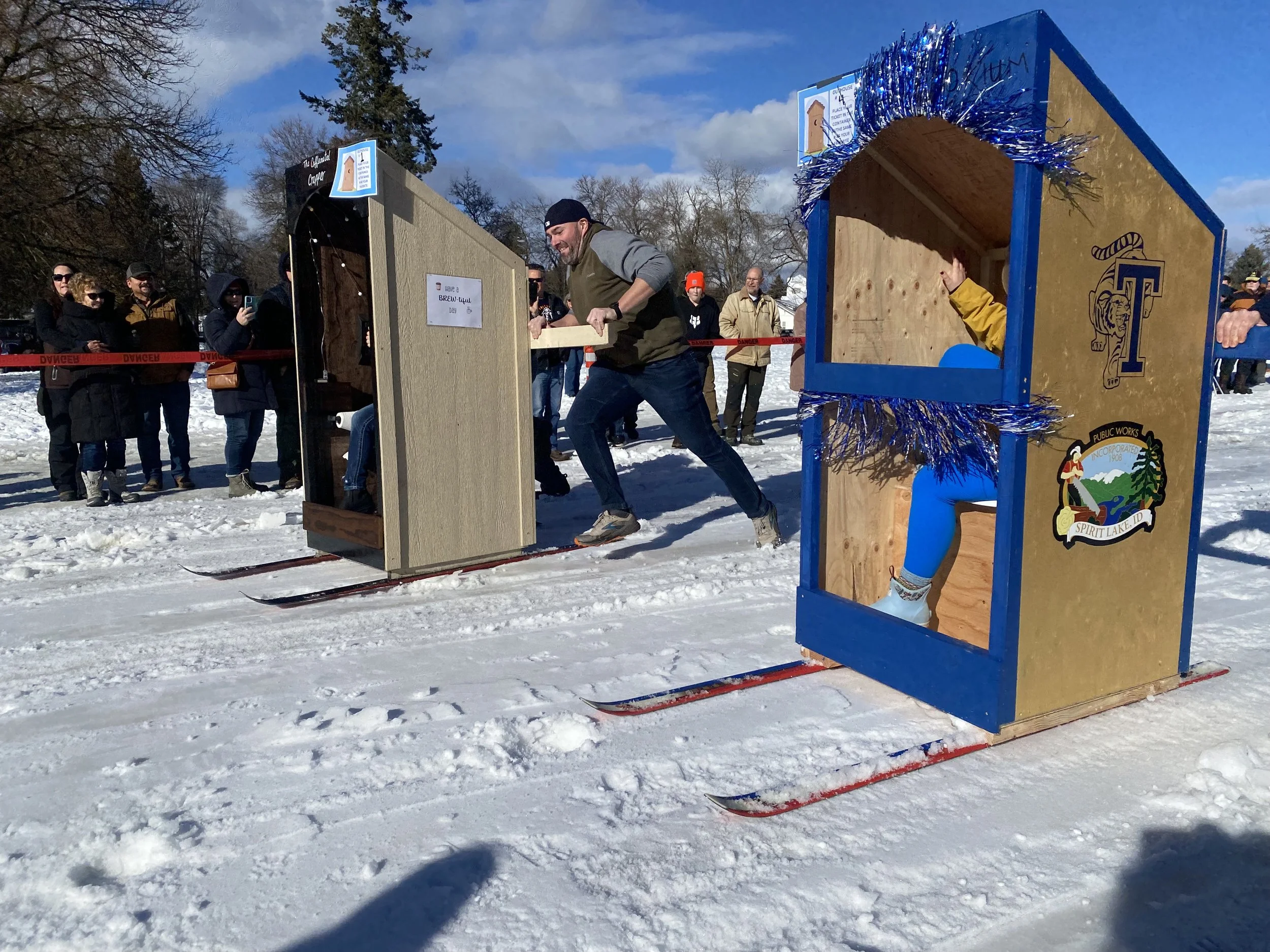 People participating in a fun outdoor event on snow, with two homemade cardboard sleds, one with a toilet bowl design and the other decorated with blue tinsel, and an audience watching nearby.