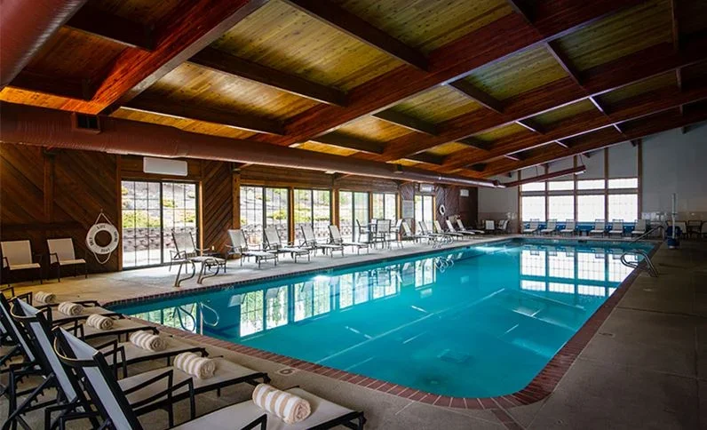 Indoor swimming pool area with lounge chairs and large windows showing outdoor scenery.