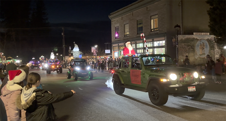 Crowd of people on a street decorated with Christmas lights and brightly lit buildings during a Christmas parade at night.