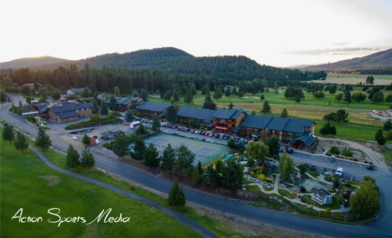 Aerial view of a large hotel or resort with parking and tennis courts, surrounded by trees and mountains.