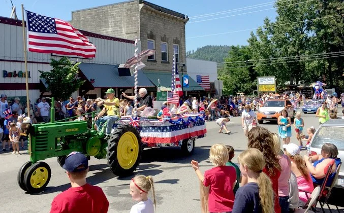 A parade with a decorated float pulled by a green tractor, with American flags and patriotic decorations, moving through a small town street lined with spectators of all ages celebrating Independence Day.