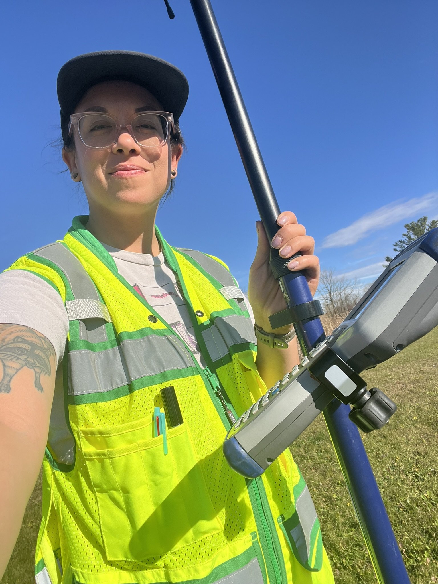 A person wearing safety glasses, a baseball cap, a yellow reflective vest, and a gray uniform, holding a surveying instrument outdoors on a sunny day with a blue sky and some trees in the background.