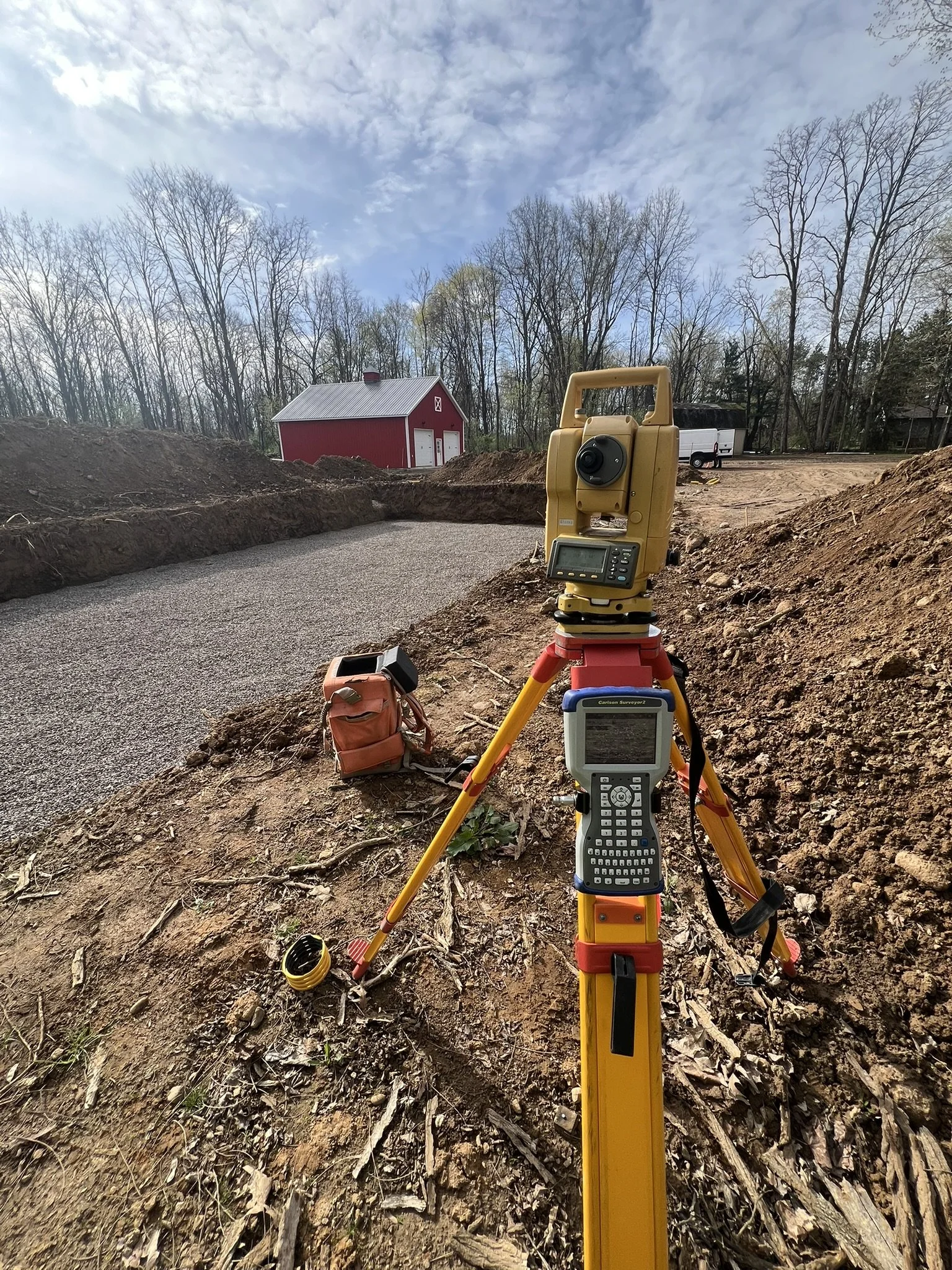 Surveying equipment set up on a dirt construction site with a red barn, a white van, and a wooded background under a partly cloudy sky.