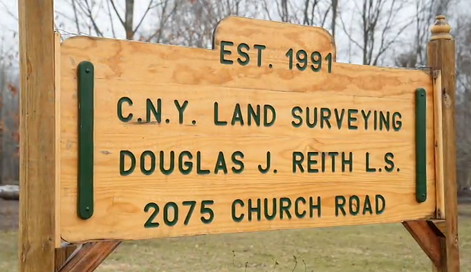 Wooden sign with green letters that reads "Est. 1991 C.N.Y. Land Surveying Douglas J. Reith L.S. 2075 Church Road" against a background of bare trees.