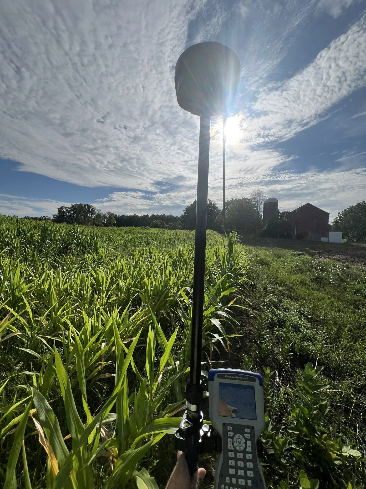 A person holding a surveying instrument in a lush green field with farm buildings and trees in the background under a partly cloudy sky, with the sun shining brightly.