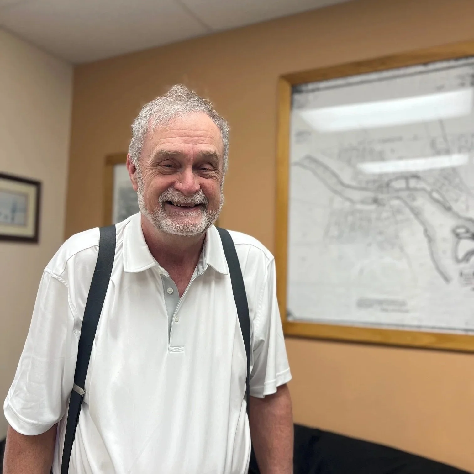 An older man with gray hair and a beard, smiling, wearing a white polo shirt and suspenders, standing in an indoor setting with a framed picture on the wall and a map in the background.