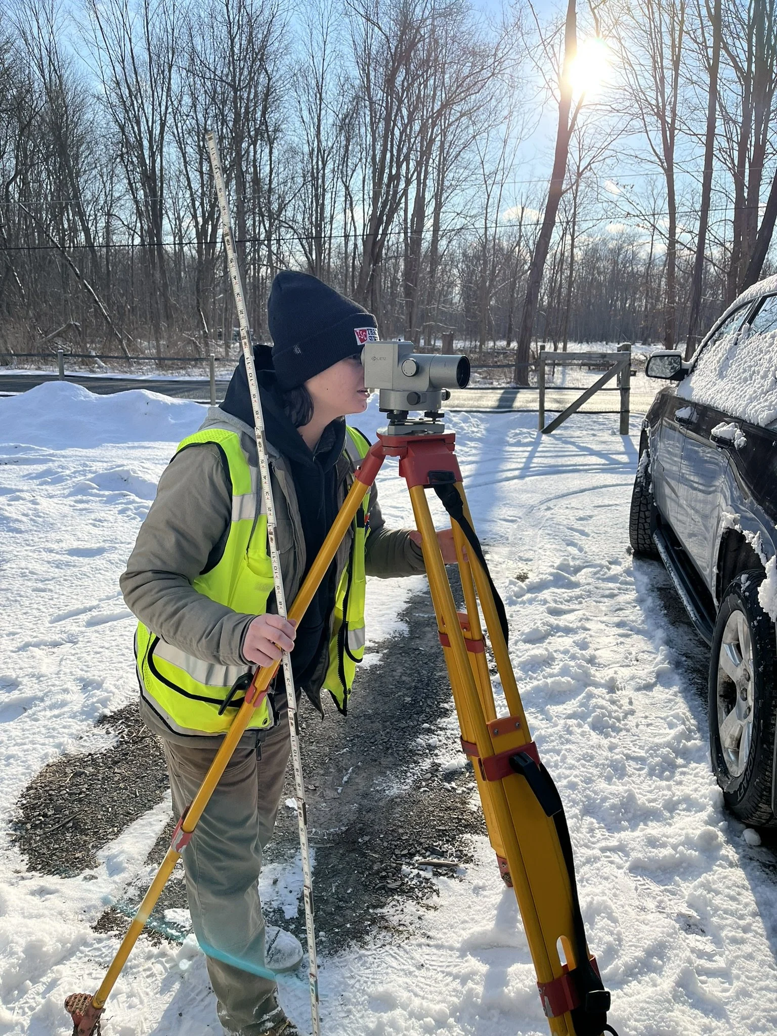 A person in winter clothing using a surveying instrument on a tripod in a snowy outdoor setting with trees and a car covered in snow.