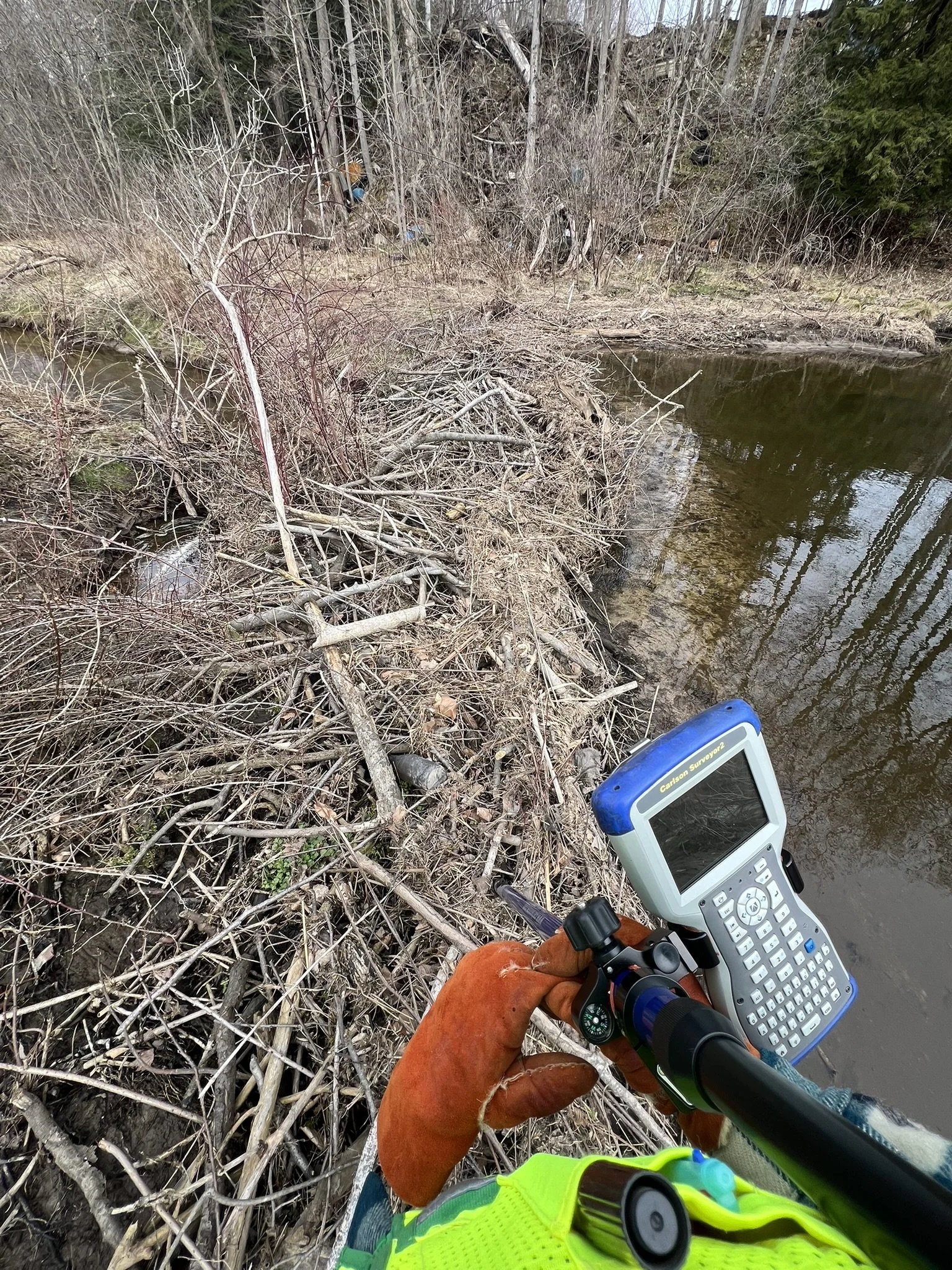A person wearing orange gloves and a neon yellow safety vest holds a handheld GPS or survey device, standing near the edge of a river or creek with dry branches and twigs along the bank, and trees in the background.
