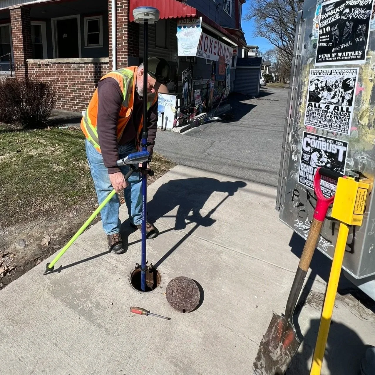 A man in an orange safety vest is working on a sewer cover on a sidewalk, using a drain cleaning machine. There is a small screwdriver on the ground nearby, and construction tools are visible, including a shovel with a red handle and a yellow safety box. The scene is outdoors, next to a brick building with small bushes and a street in the background.