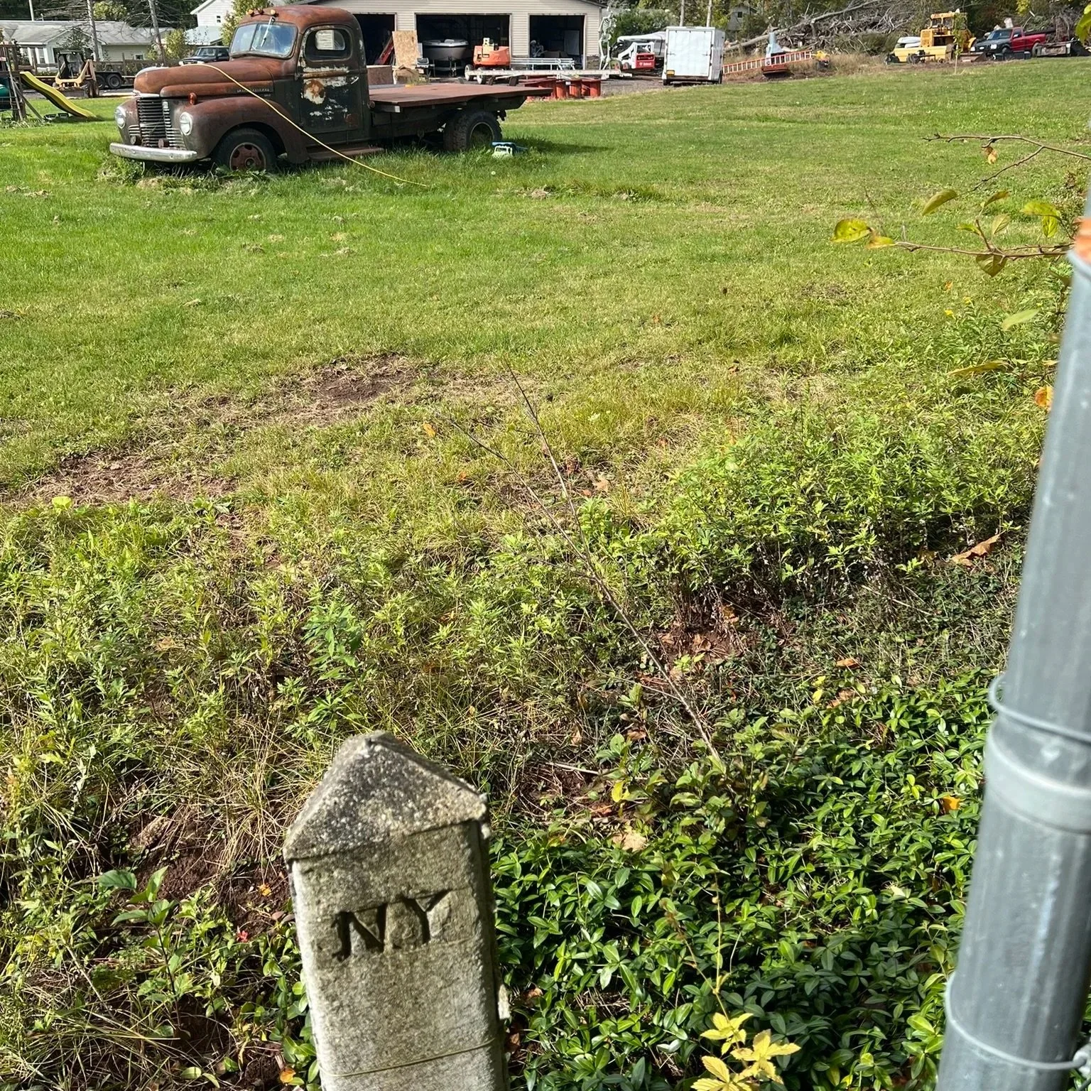 Old rusted pickup truck parked on a grassy lawn with a house and various equipment in the background.