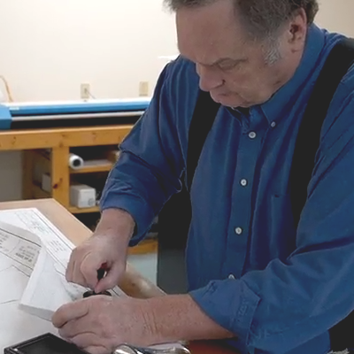 A man with gray hair in a blue shirt looking down, working on a paper at a workbench.
