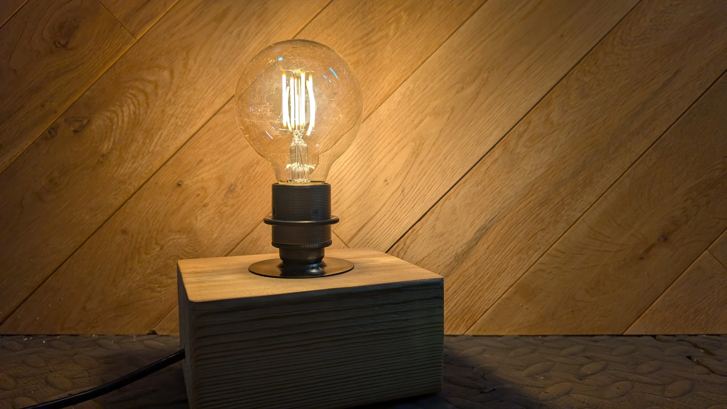 A glowing vintage-style light bulb with an exposed filament sits on a wooden base against a diagonal wooden wall.