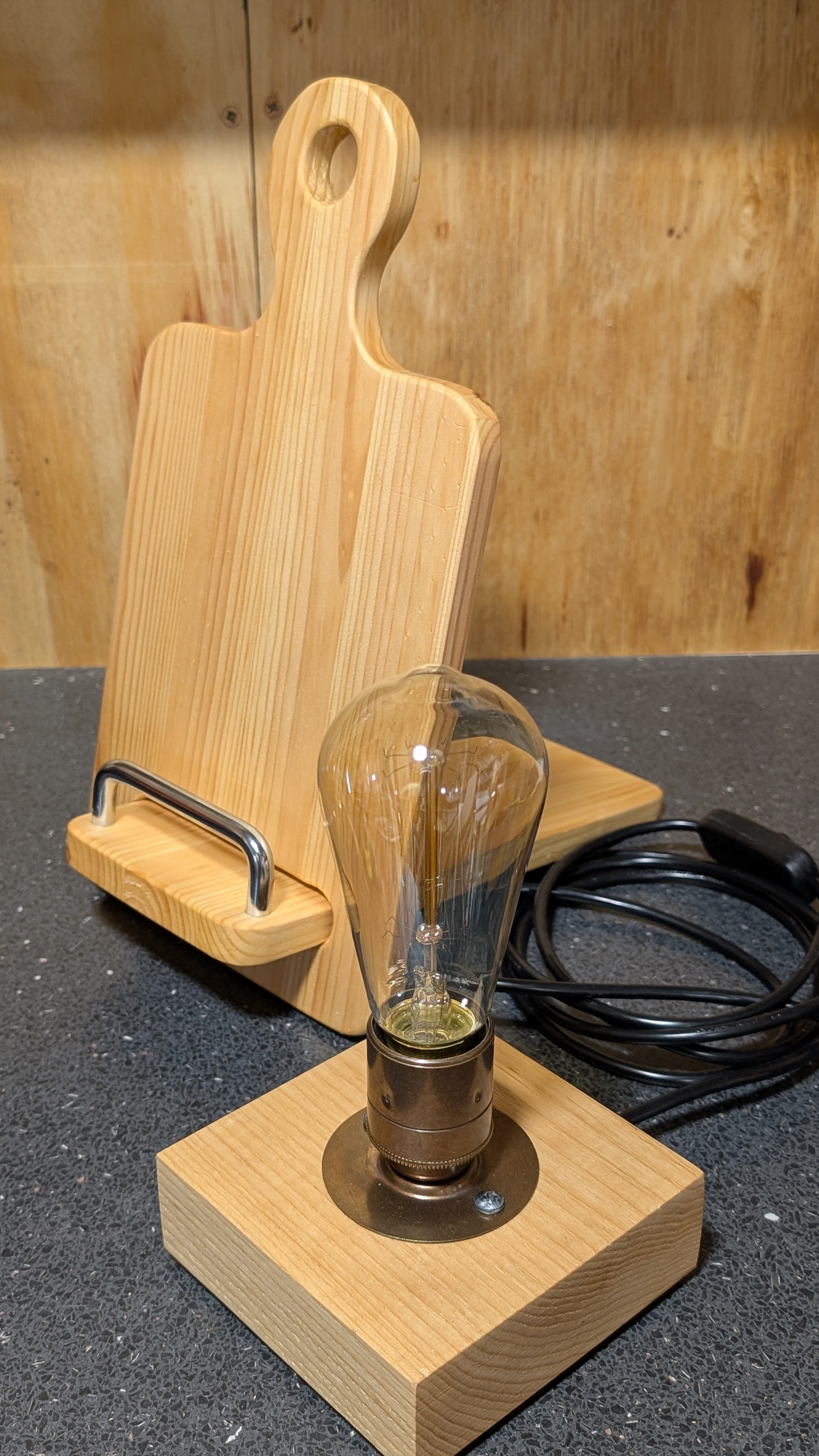 A decorative table lamp with an exposed filament bulb, on a wooden base, with a wooden cutting board/ menu stand.