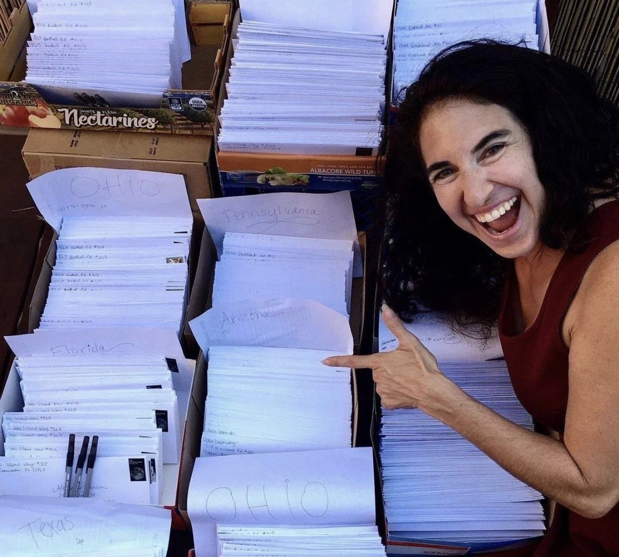 A woman smiles and points to a large box of prepared letters.