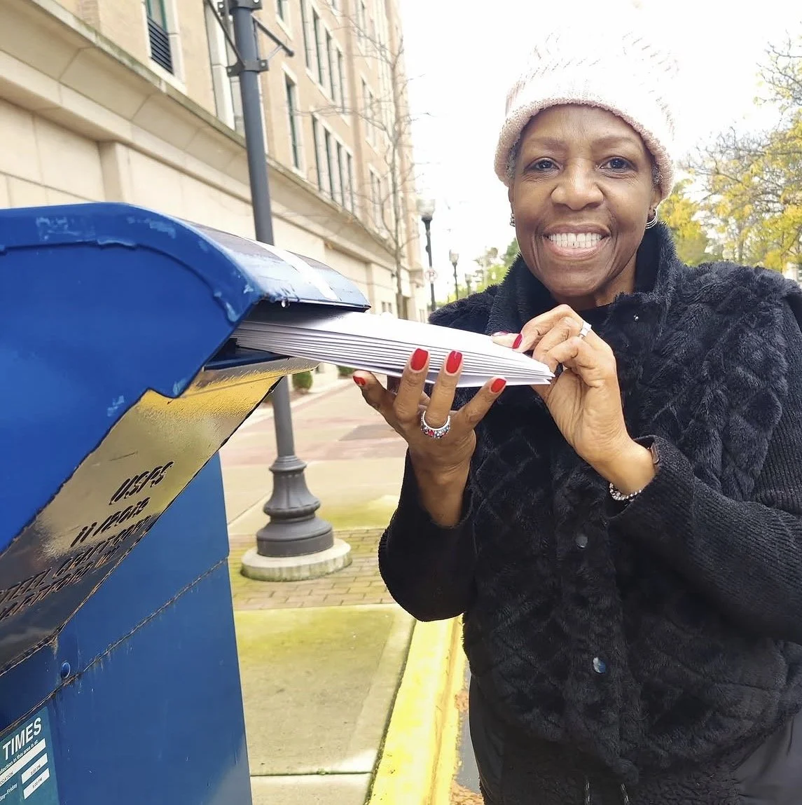 A woman in a hat placing a stack of Vote Forward letters in a a blue USPS mailbox