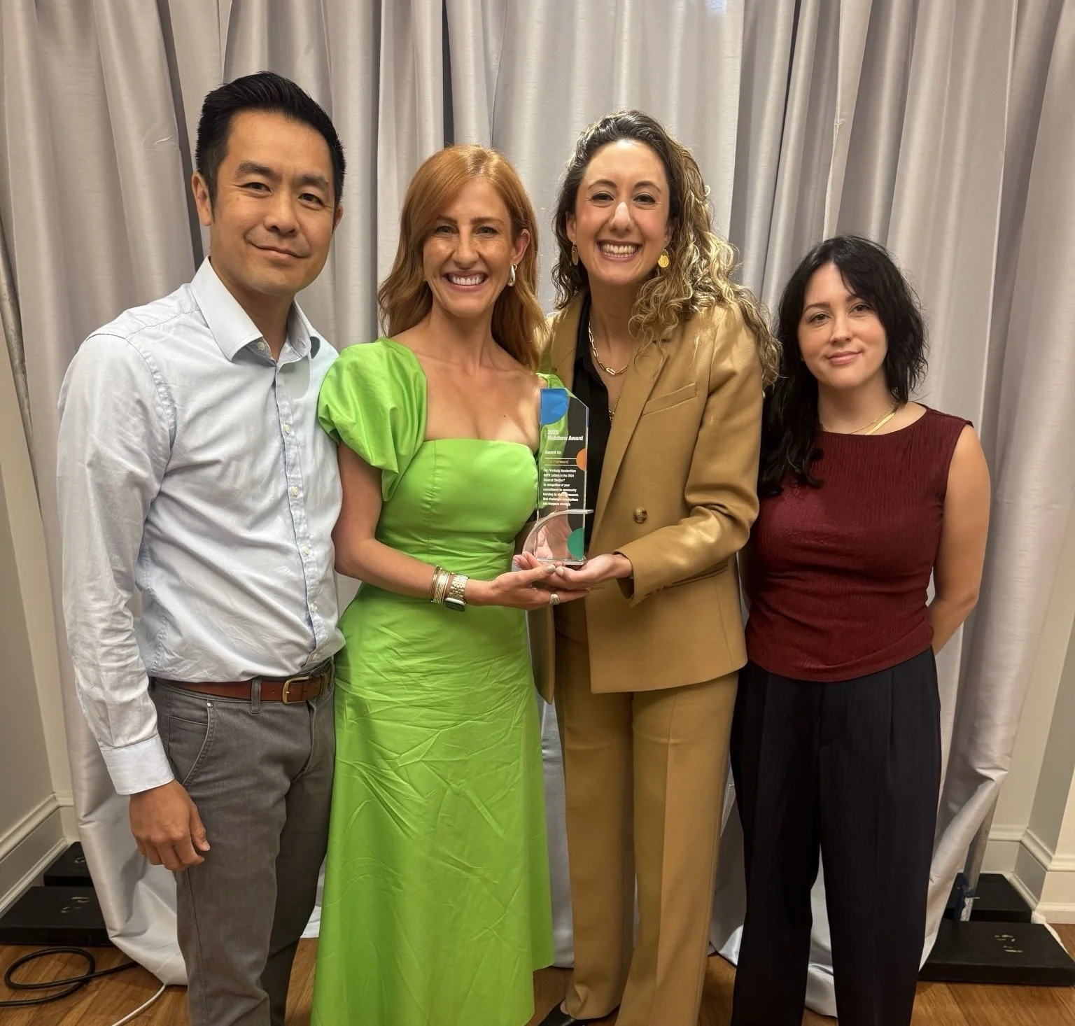 Four people standing together, smiling, with one woman holding a trophy or award in the center, in front of a curtain backdrop.