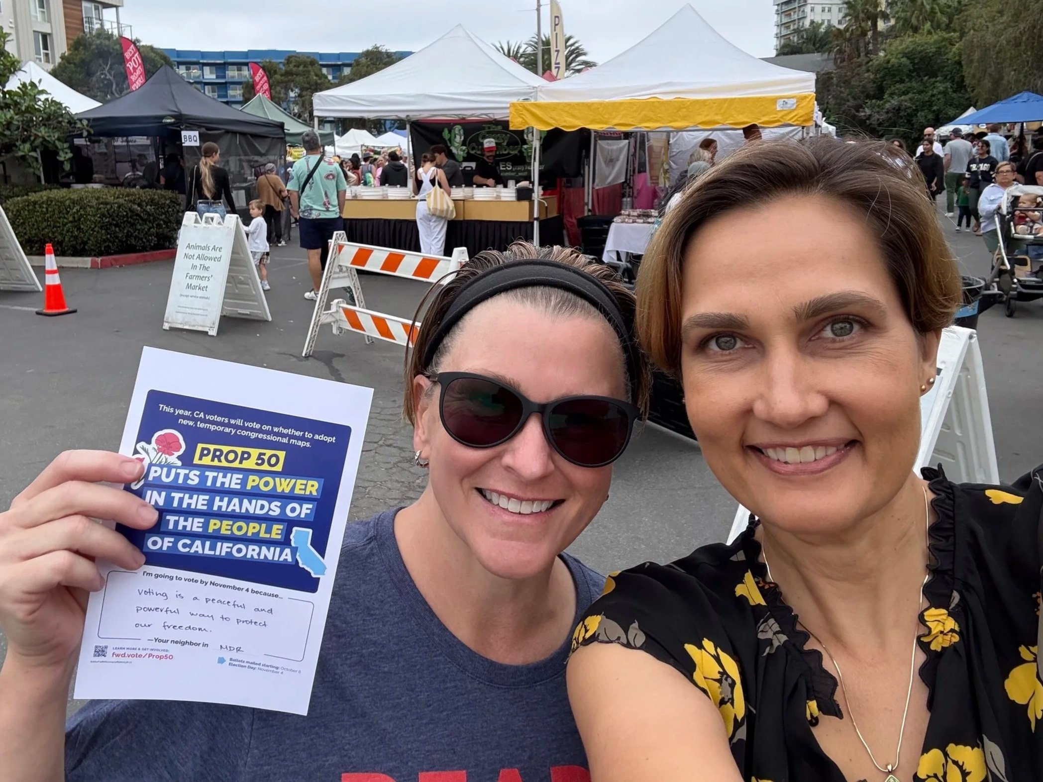 Two people at a farmers market. The person on the left is smiling and holding up a flyer about the CA Prop 50 election.
