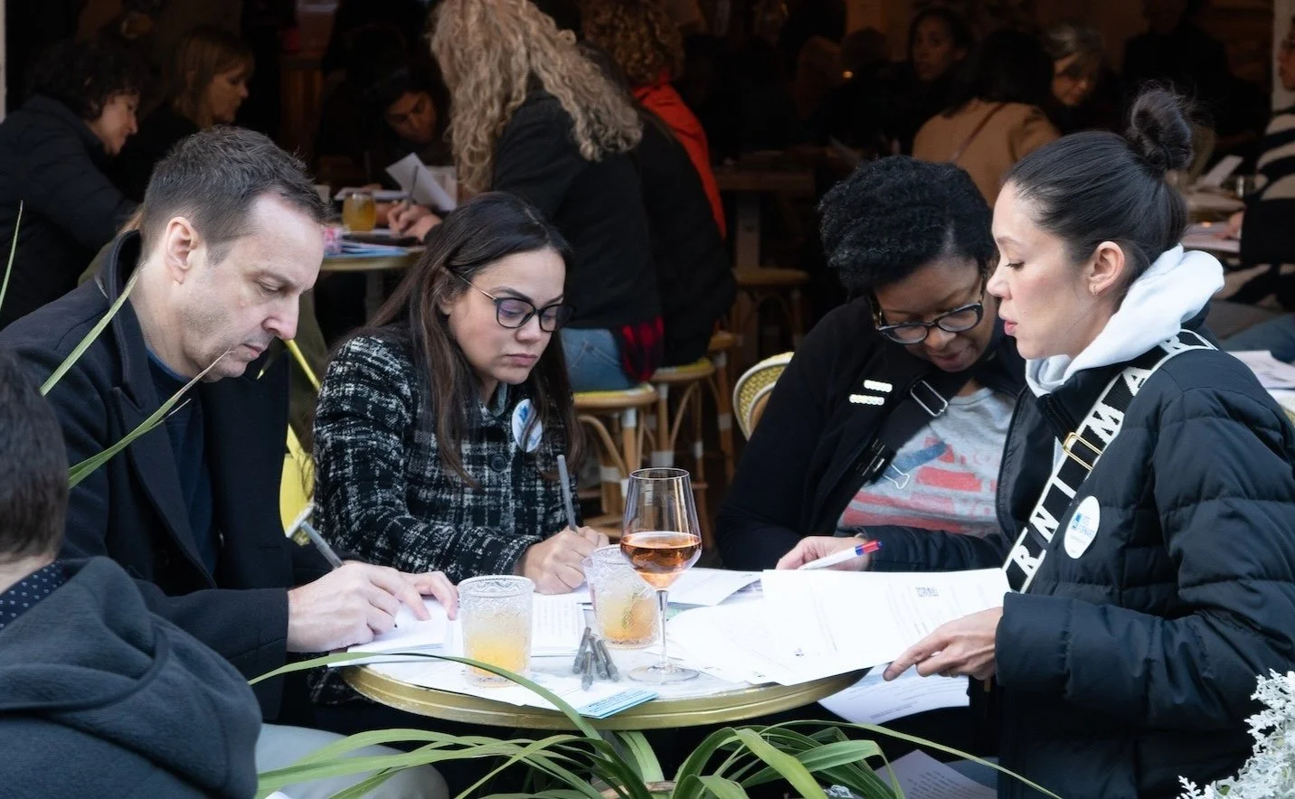 Four people sit at a table drinking wine and beer and writing letters.
