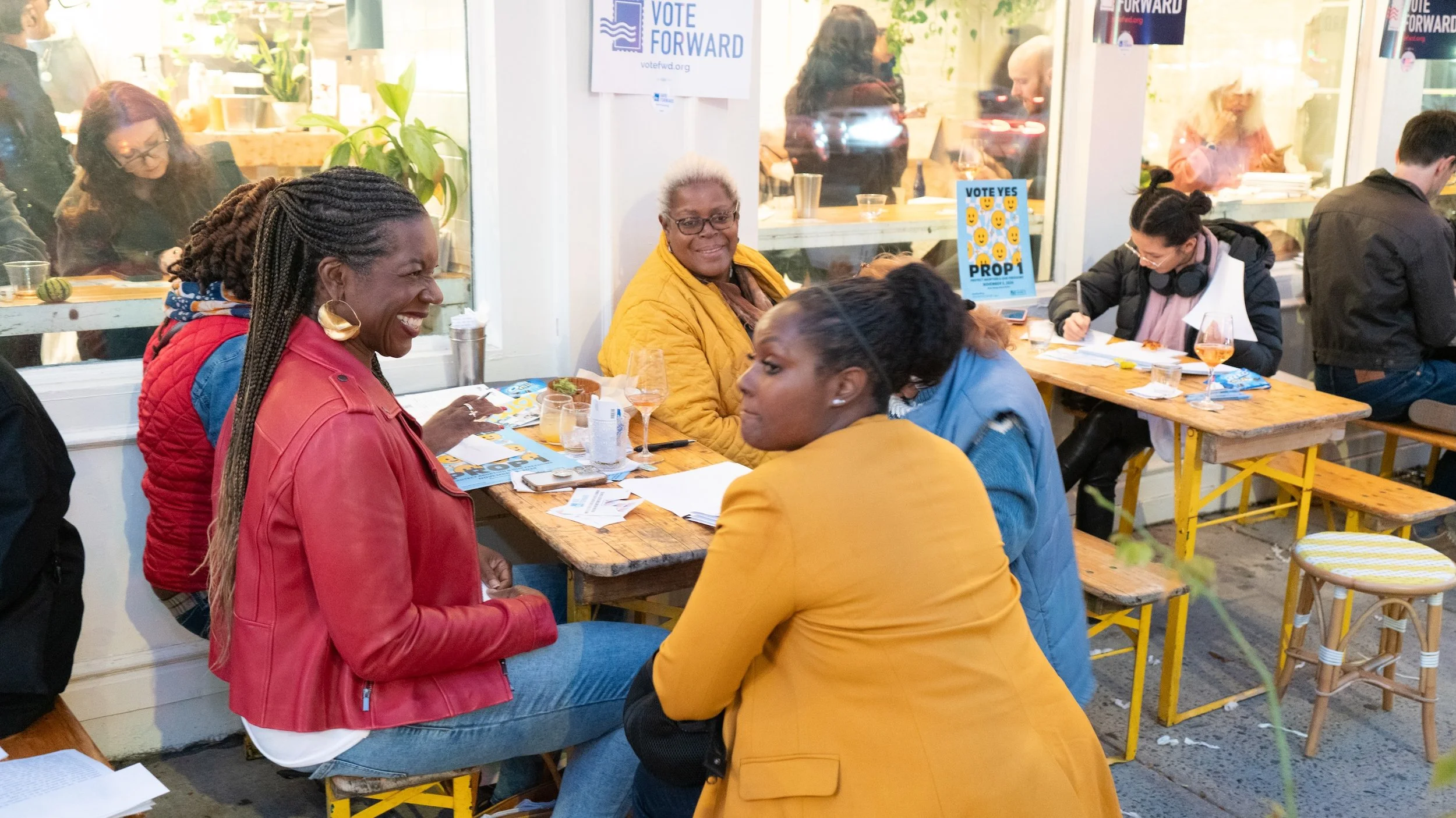 A group of women sit outside around a table while writing letters.