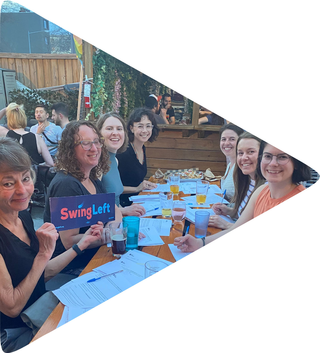 A group of people sitting on an outdoor bar patio at a table full of drinks and letters. A woman in front is holding up a Swing Left placard.