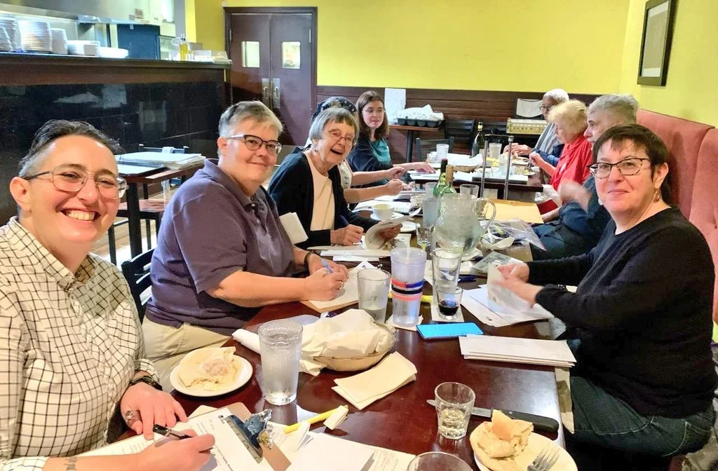 A group of people sit around a restaurant table smiling and writing letters.