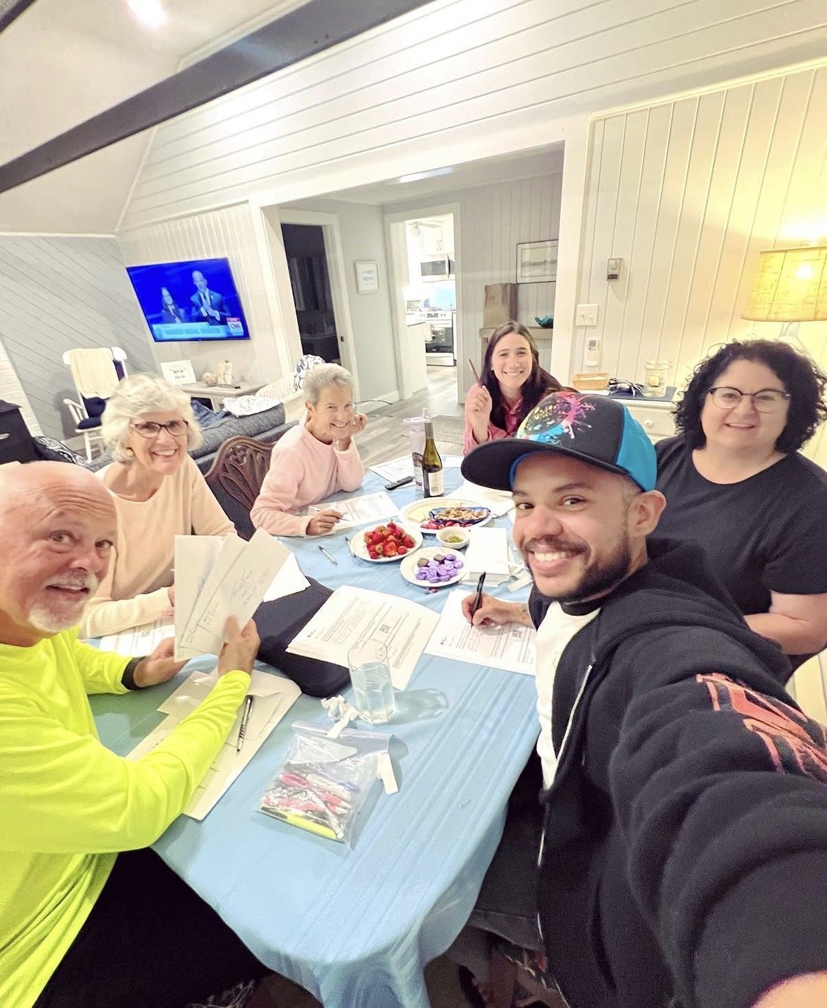 A group of six smiling people sit around a table at home eating food and writing letters.