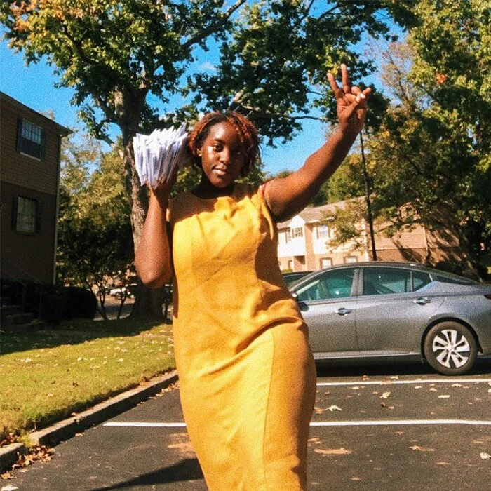 A woman in a yellow dress flashing a peace sign with one hand and holding a stack of letters in the other hand.