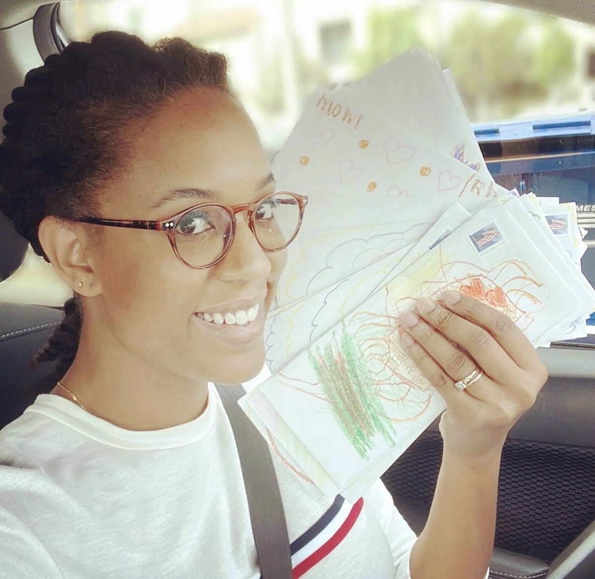 A woman with glasses sitting in her car holding up a stack of letters. The letters have children's doodles on them.