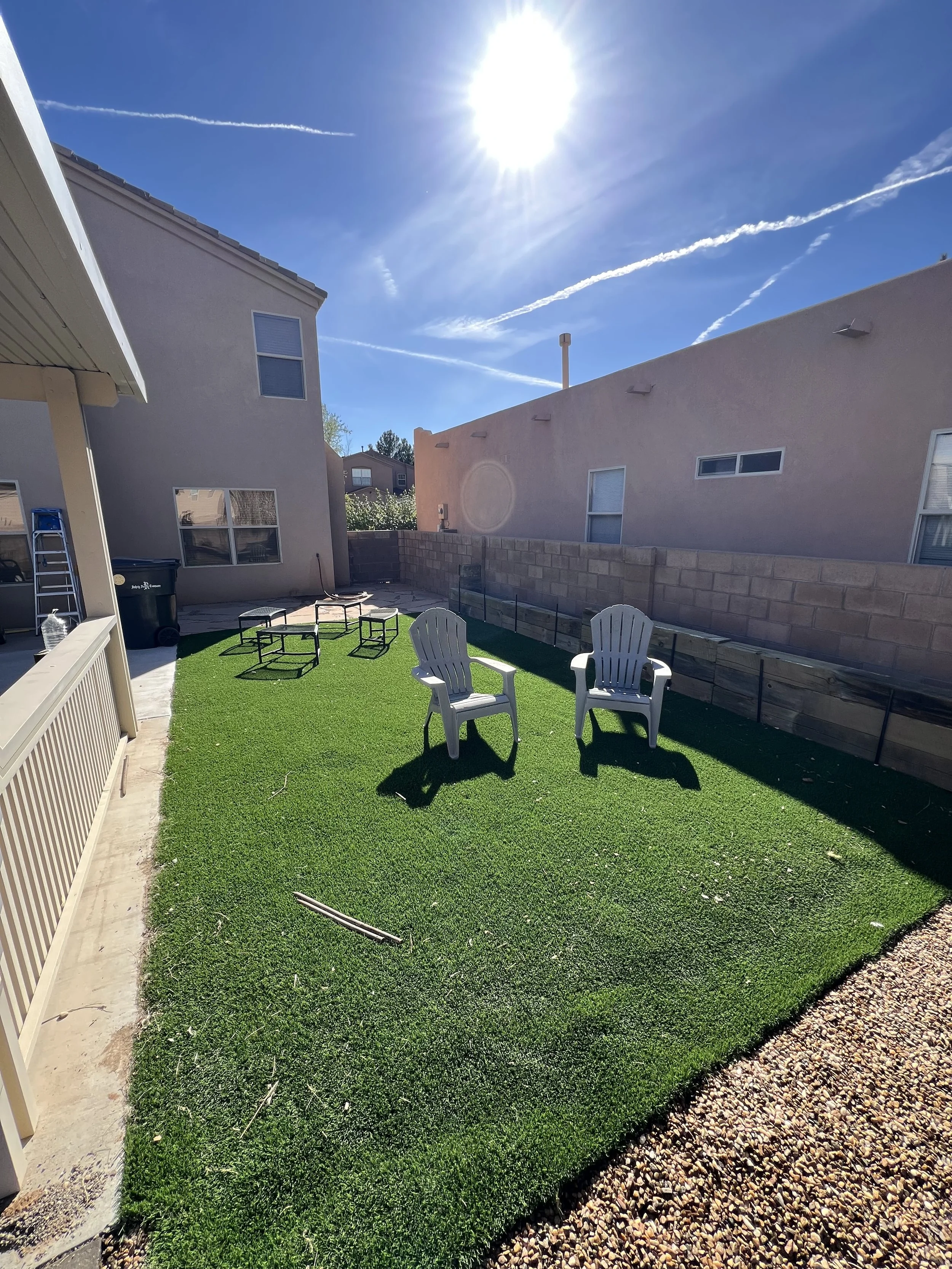 A backyard with artificial grass, two white plastic chairs, several small tables, a gravel pathway, a concrete patio, a wooden fence, and neighboring houses under a bright sunny sky.