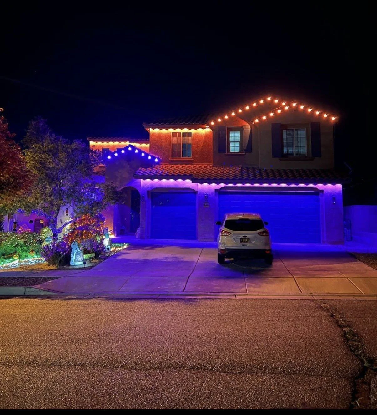A two-story house decorated with multicolored Christmas lights at night. The house has a tiled roof, two blue garage doors, and a white car parked in the driveway. The front yard has illuminated decorations and a small tree with lights.