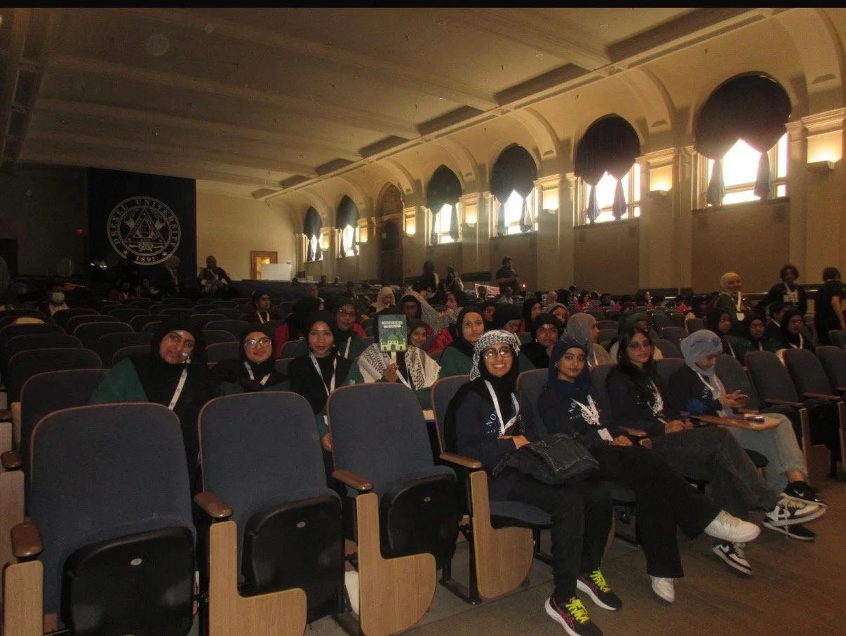 A large group of people seated in an auditorium, some smiling and facing the camera, with women wearing hijabs. The room has high ceilings and large windows with draped curtains, and a black backdrop with a seal or emblem on the stage.