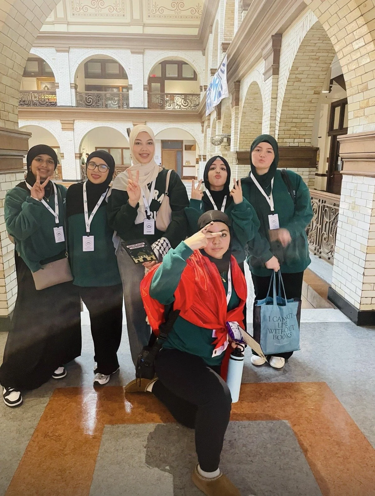 Group of six young women, some wearing hijabs, posing inside a building with brick arches, balconies, and ornate railings. They are smiling, making peace signs, and one is saluting.