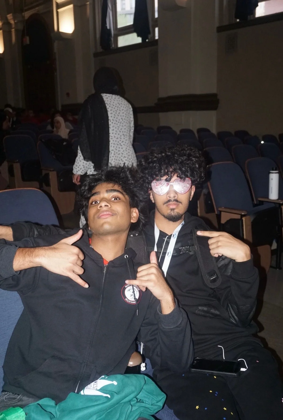 Two young men sitting in a theater, one wearing a black hoodie with a logo, making hand signs, and the other wearing sunglasses with curly hair, pointing at himself, with an auditorium filled with seats and a woman walking in the background.