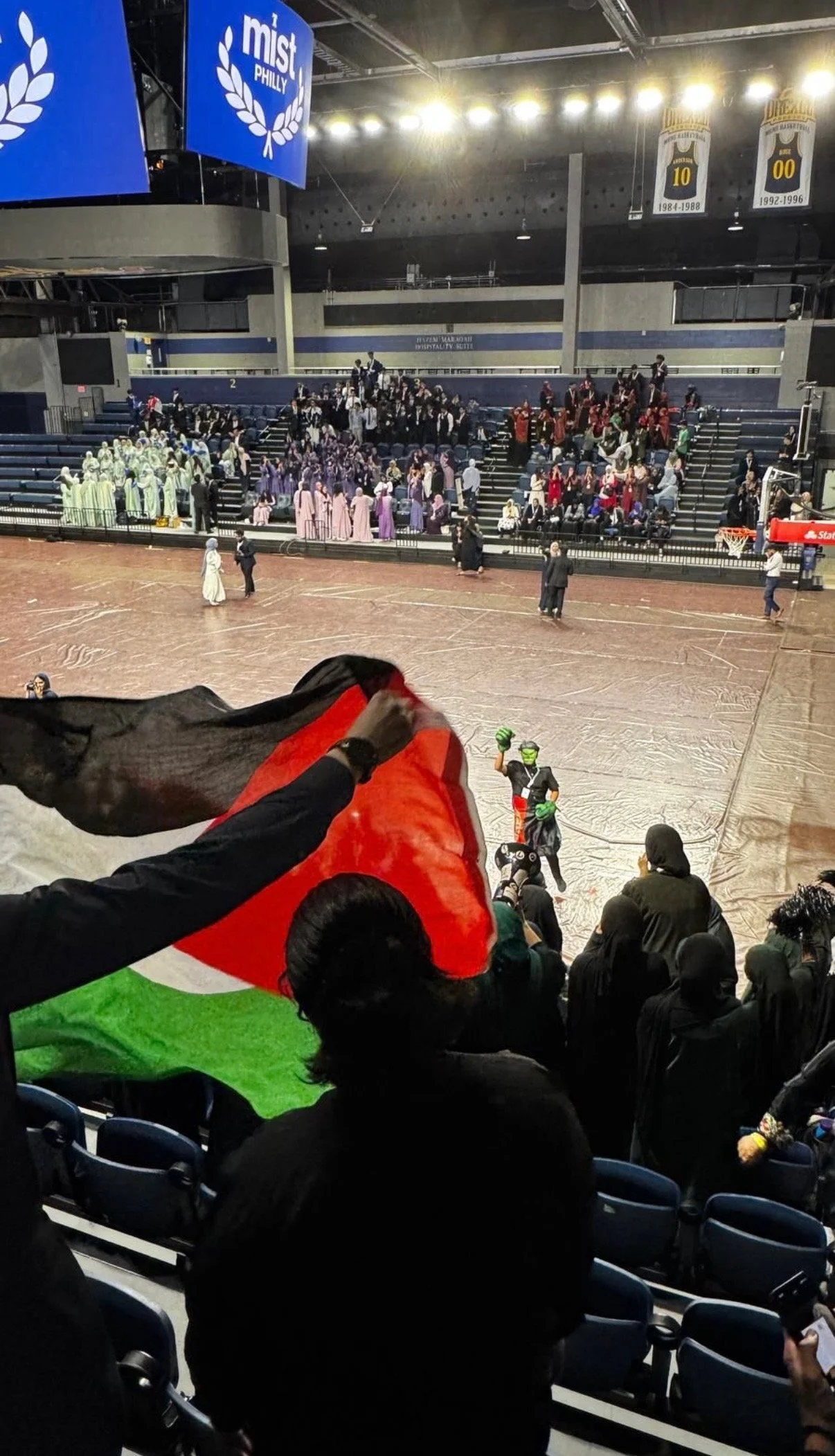 Spectators at an indoor basketball game, with some holding a large flag. The gymnasium is decorated with banners and has a group of performers dressed in colorful costumes on the court. The stands are filled with people, and a team mascot is visible 