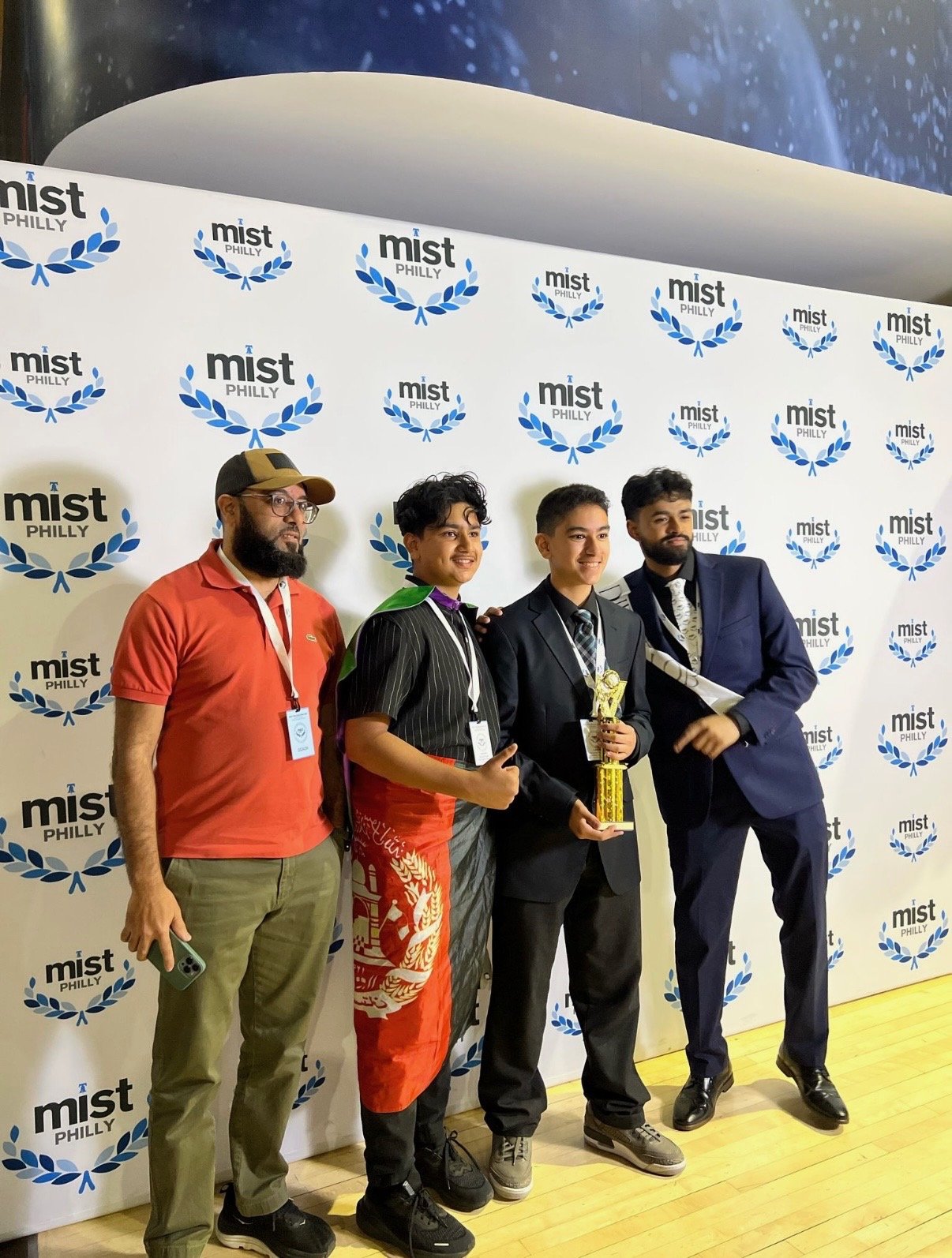 Four people standing in front of a backdrop with 'Mist Philly' logos, one holding a trophy, all dressed in formal or semi-formal attire, celebrating an event or competition.