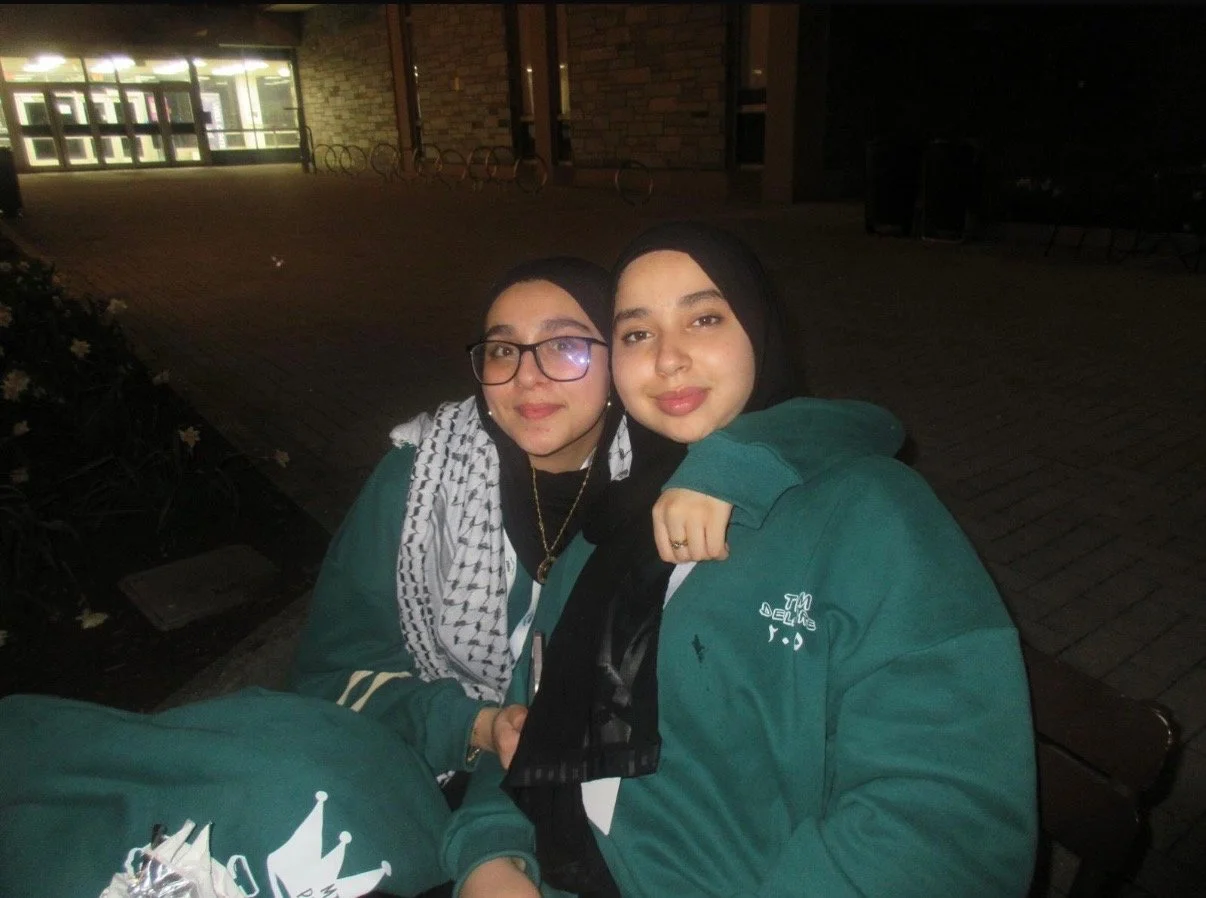 Two women sitting close together outdoors at night, one with glasses and a scarf, and the other with a black hijab, both wearing green jackets, smiling at the camera.