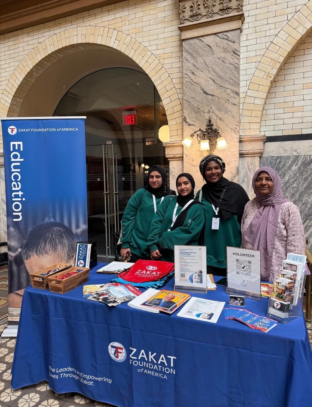 Four women and one girl standing behind a Zakat Foundation of America booth, with informational materials and volunteer sign-up sheets, inside a building with stone and brick architectural details.