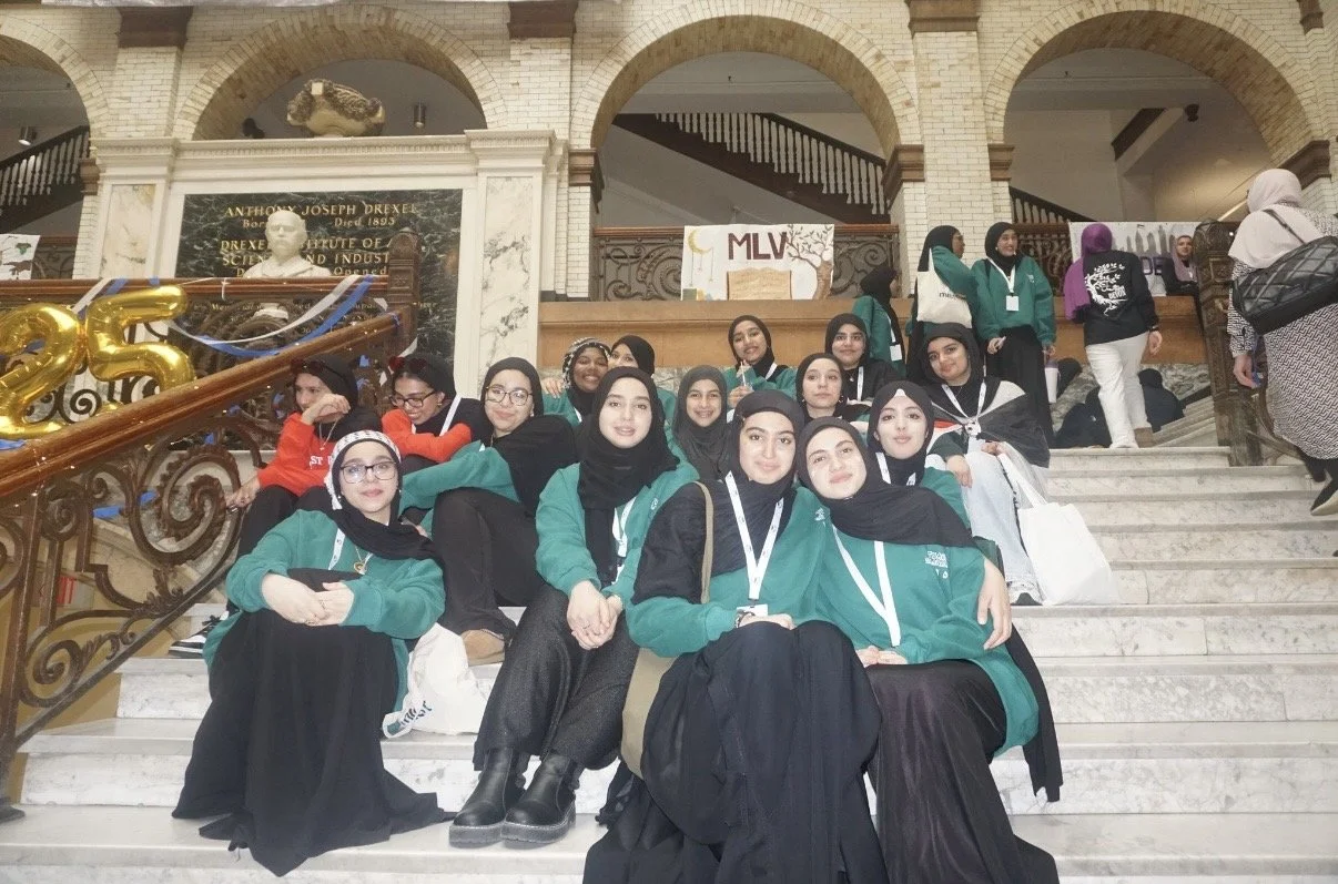 Group of young women sitting on stairs inside a historic building, possibly a museum or university, with some wearing green jackets, hijabs, and lanyards, and others wearing casual clothing, with decorations including balloons and signs in the backgr