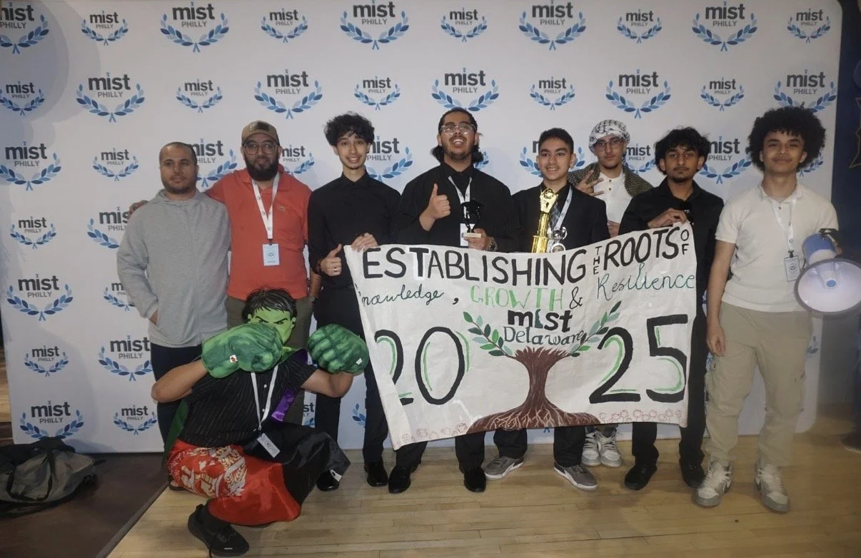 Group of young individuals holding a banner that reads 'Establishing the Roots of Knowledge, Growth & Resilience, mist Delaware, 2025' at a Mist Philly event. One person is dressed as a superhero with green gloves, and some are wearing lanyards and b