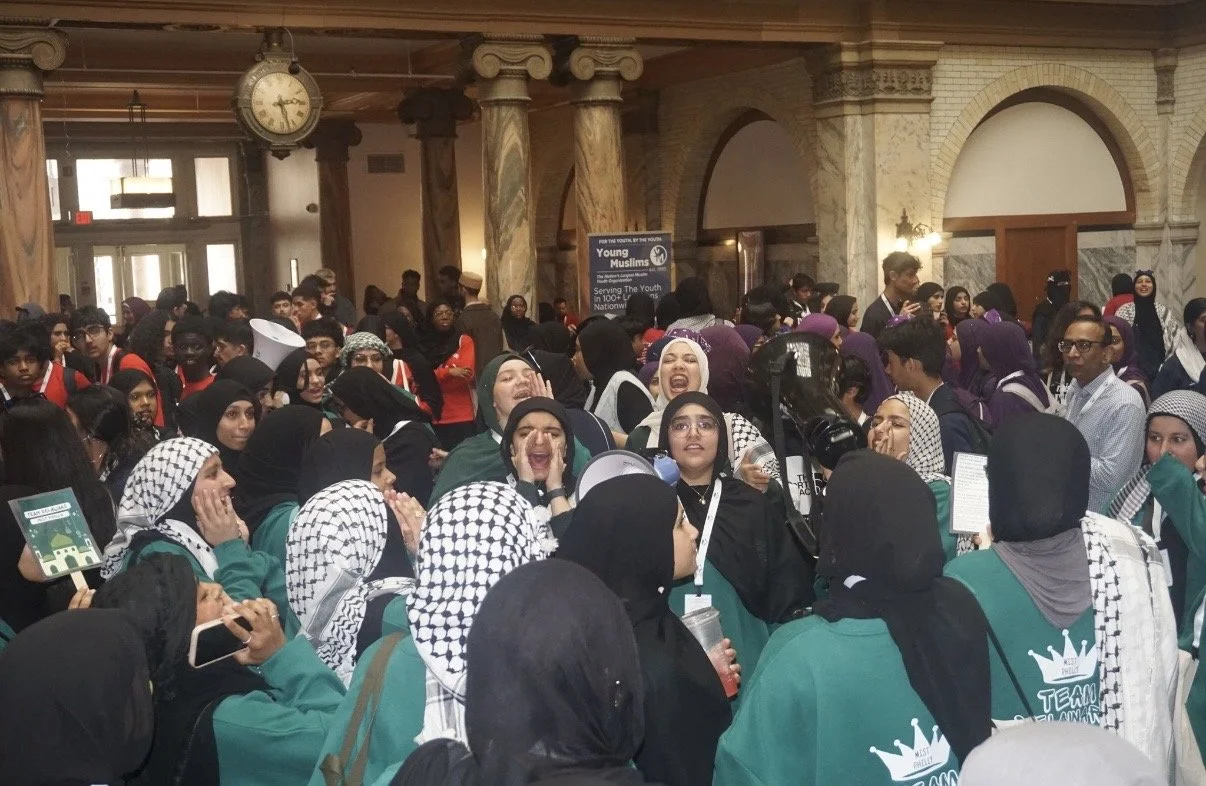 A large group of young Muslim women and men gathering indoors, many wearing headscarves and teal shirts with a crown logo. The scene appears lively and joyful, with some smiling and laughing.