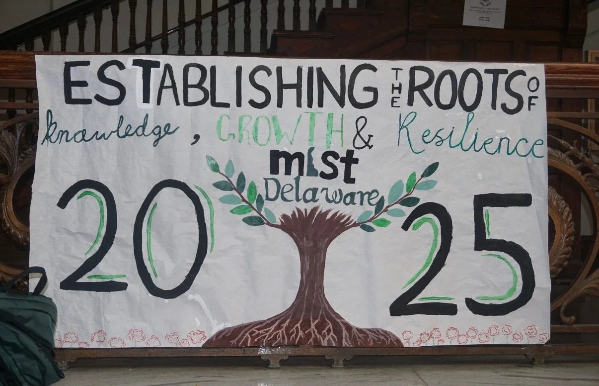 A hand-painted banner with the message 'Establishing the roots of knowledge, growth, and resilience must delaware 2025.' The banner features a tree with roots and leaves in the center, and a decorative border at the bottom.