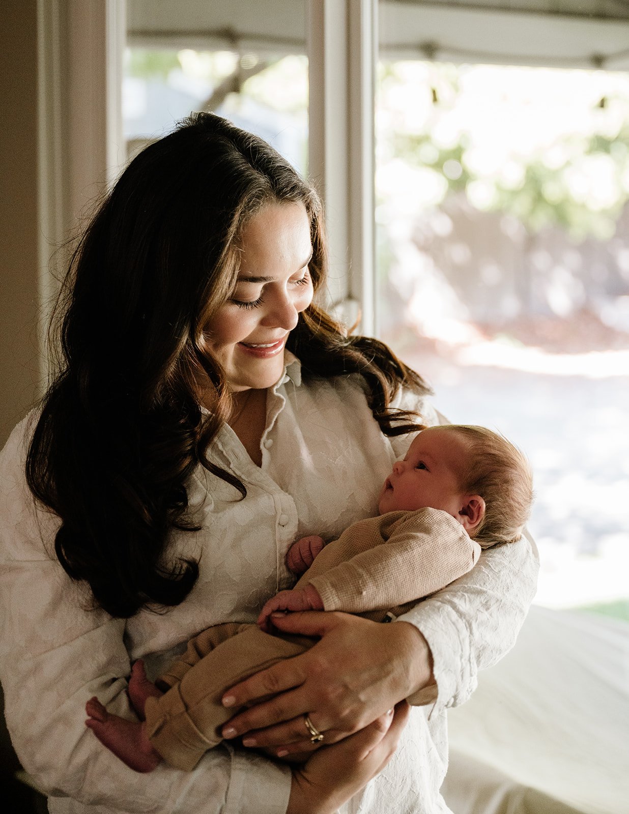 A woman holding a baby indoors near a window, smiling and looking at the infant, who is dressed in beige clothing.