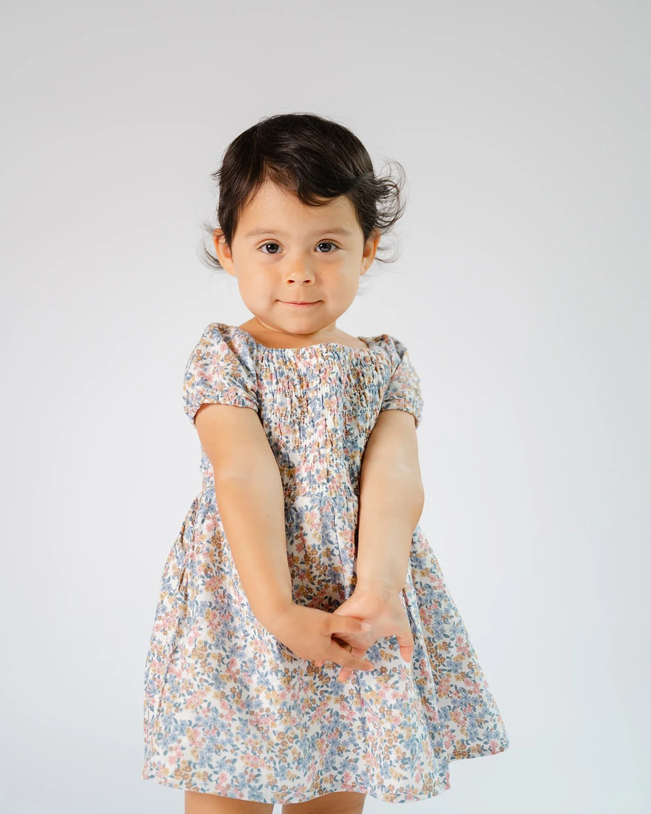 A young girl with dark hair and big eyes wearing a floral dress stands with arms crossed in front of a plain, light gray background.