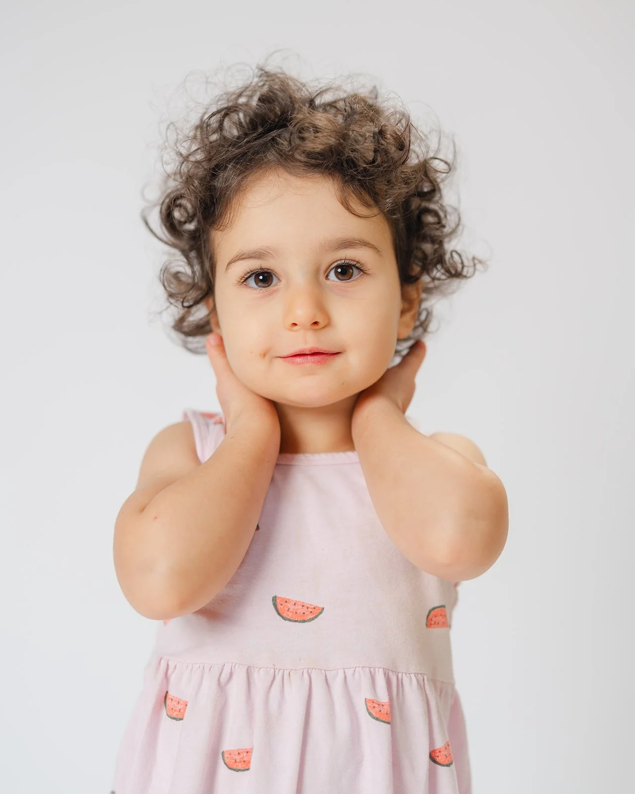 A young girl with curly brown hair and light skin, wearing a pink dress with watermelon prints, holding her hands behind her neck, looking at the camera with a neutral expression.