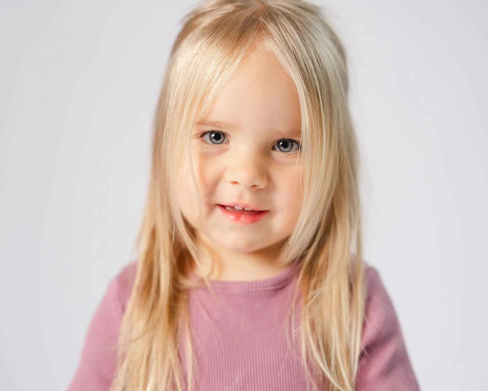 A young girl with long blonde hair and blue eyes wearing a mauve sweater standing against a plain white background.