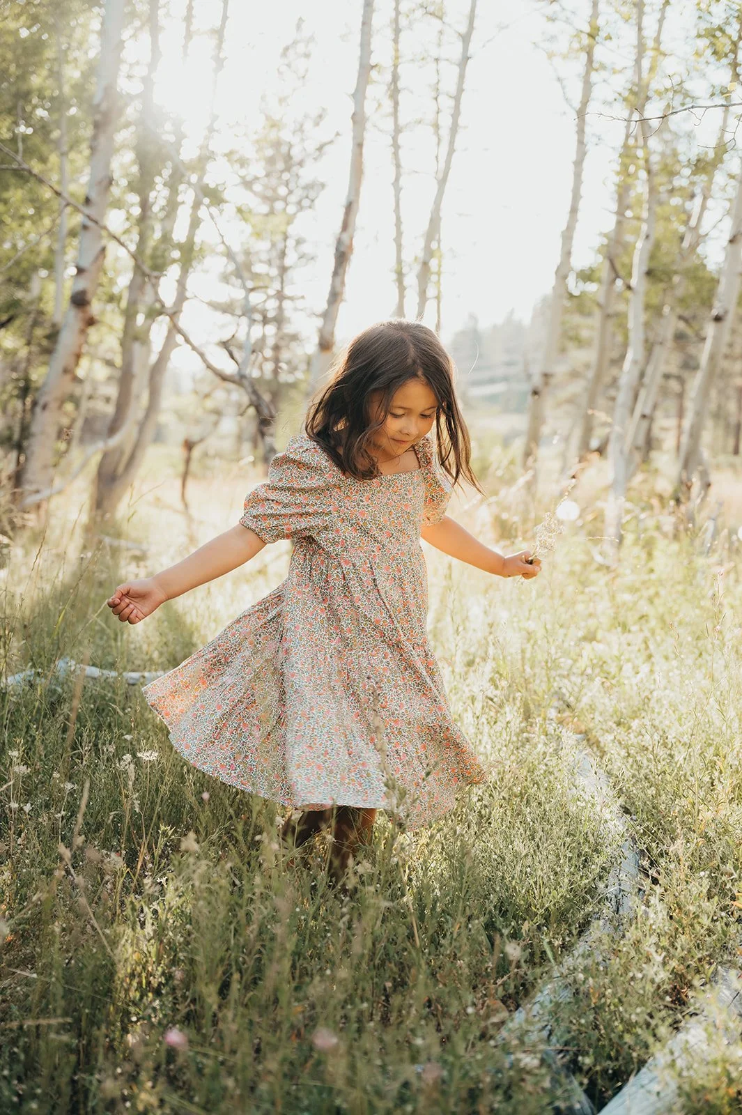 A young girl in a floral dress spinning in a grassy field surrounded by trees, bathed in sunlight.