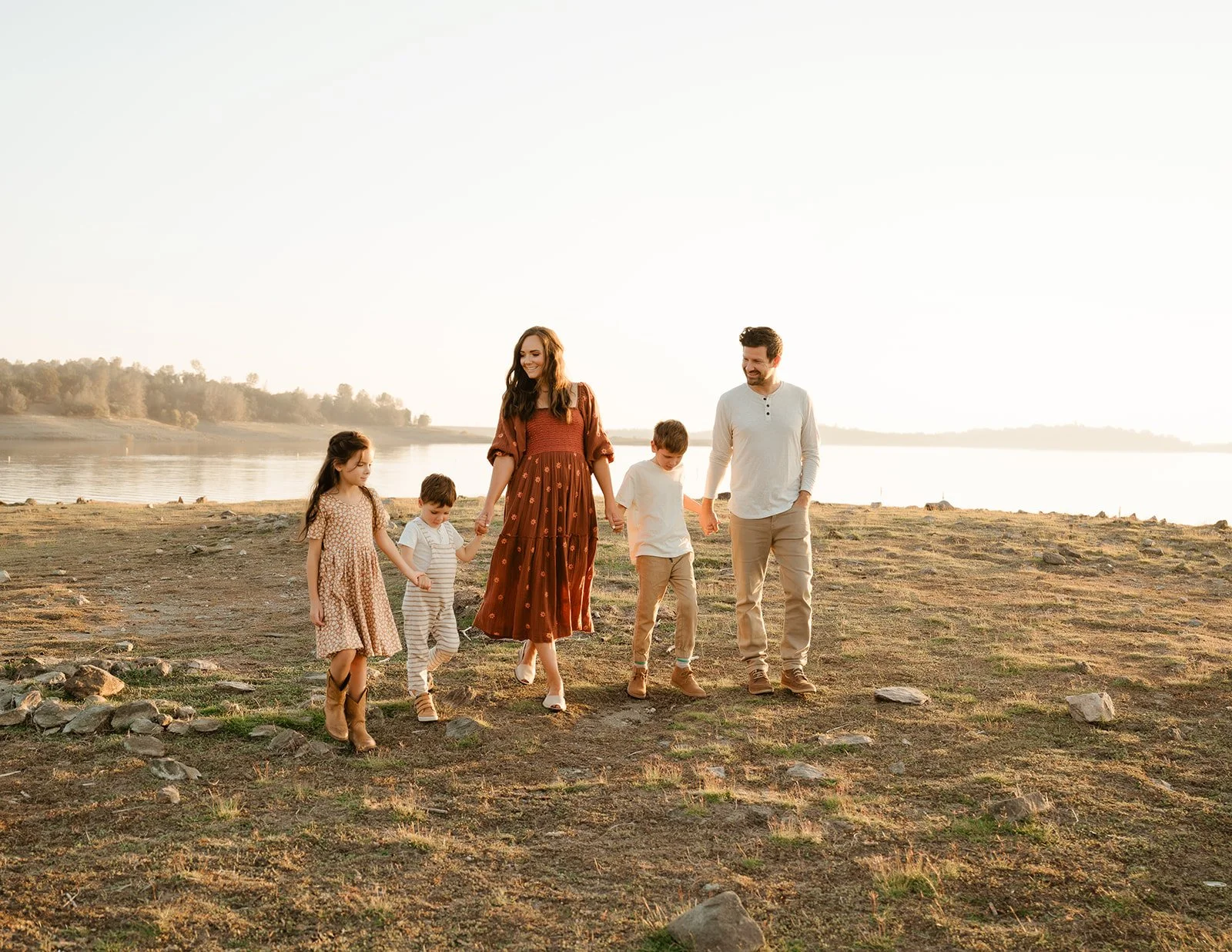 A family of six walking hand in hand along a lakeside during sunset, with a woman in a long red dress, three children, and a man in casual clothing.