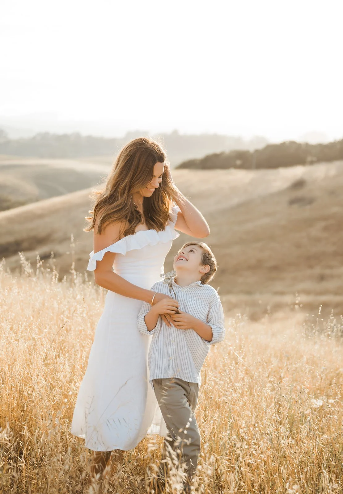 A woman and a young boy walking through a field of tall, golden grass during sunset, smiling and looking at each other.